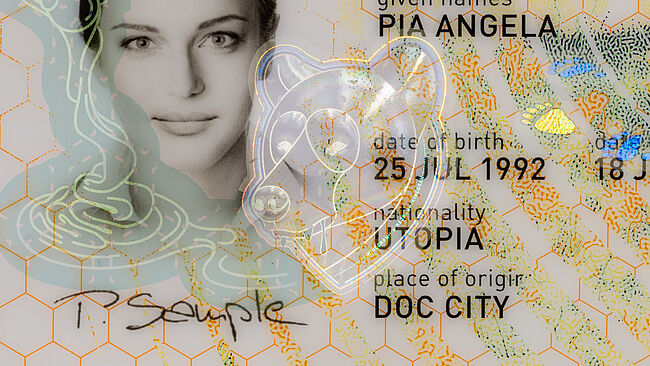 Close-up image of sample passport data page with a KINEGRAM PCI with Full Data Protection security feature whose motifs show a bear stealing honey from a beehive. The close-up focuses on the bear licking up spilled honey.